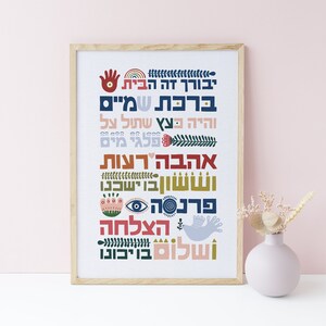 May include: A framed print with a white background and colorful Hebrew text. The text includes the words "Blessed is this home", "Blessings from the heavens", "May you be like a tree planted by the waters", "Love and kindness", "Light and joy", "Peace and prosperity", "Success and good fortune", and "Shalom". The print is decorated with various symbols, including a hand, a rainbow, a tree, a heart, a menorah, an eye, a flower, a dove, and a star.