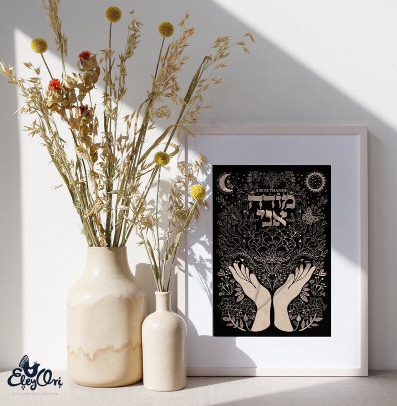 May include: A black and gold print with the Hebrew words "Todah Rabah" meaning "I give thanks" in a decorative floral design with hands raised in prayer. The print is framed in a white frame and is displayed on a table with dried flowers in a vase.