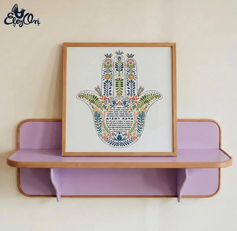 K&ouml;nnte beinhalten: Ein gerahmter Hamsa-Hand-Kunstdruck mit farbenfrohen Blumenmustern, der auf einem lilafarbenen Regal mit Holzrahmen ausgestellt ist. Das Kunstwerk zeigt hebr&auml;ischen Text und ist vor einer cremefarbenen Wand platziert.