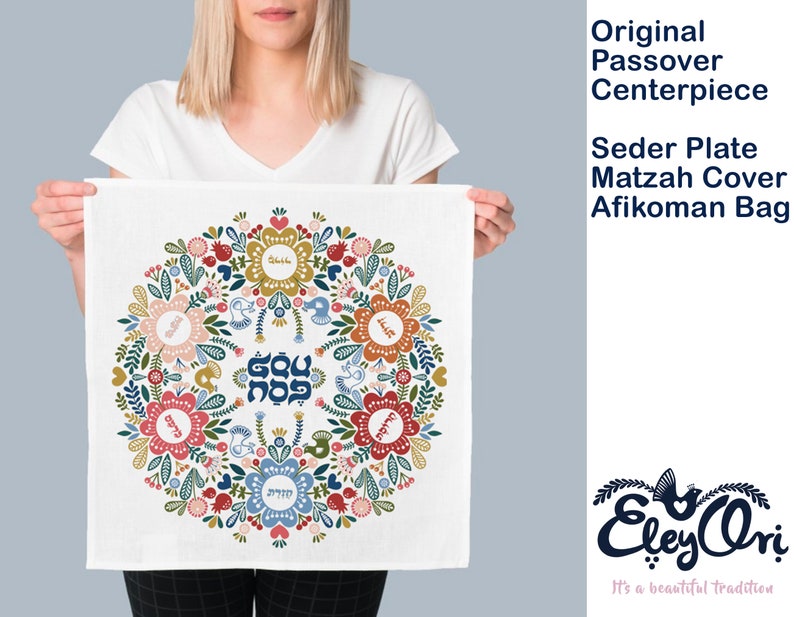 May include: A white fabric Passover Seder plate cover with a colorful floral design and Hebrew text. The design features a circle of flowers with a central star and the words "Dayenu" and "Ma Nishtana" in Hebrew.