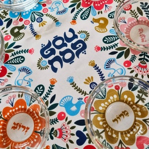 May include: A white tablecloth with a floral pattern and Hebrew text. Five clear glass bowls are arranged on the tablecloth. The bowls have a floral design and Hebrew text.