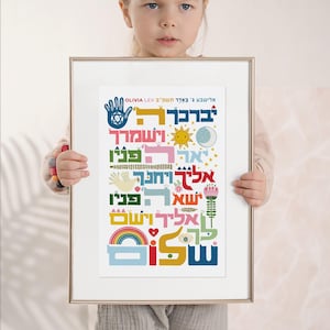 Puede incluir: Un cartel enmarcado con texto hebreo colorido, sostenido por un ni&ntilde;o. El texto incluye varios s&iacute;mbolos como una mano con una Estrella de David, un sol, una luna, un arco&iacute;ris y flores. El marco es dorado.