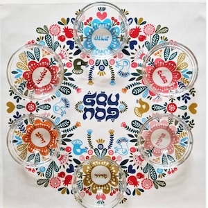 May include: A white tablecloth with a colorful floral pattern and six clear glass bowls with floral designs. The bowls are arranged in a circle around a blue and white centerpiece with the Hebrew words "L'chaim" written on it.