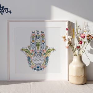 K&ouml;nnte beinhalten: Gerahmter Kunstdruck mit einem farbenfrohen Hamsa-Hand-Design mit floralen und botanischen Elementen. Das Kunstwerk enth&auml;lt hebr&auml;ischen Text und wird in einem wei&szlig;en Rahmen pr&auml;sentiert. Eine Vase mit Trockenblumen steht neben dem gerahmten Druck.