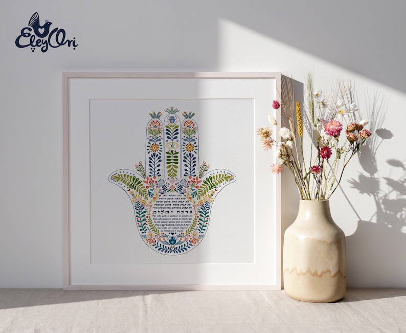 K&ouml;nnte beinhalten: Gerahmter Kunstdruck mit einem farbenfrohen Hamsa-Hand-Design mit floralen und botanischen Elementen. Das Kunstwerk enth&auml;lt hebr&auml;ischen Text und wird in einem wei&szlig;en Rahmen pr&auml;sentiert. Eine Vase mit Trockenblumen steht neben dem gerahmten Druck.