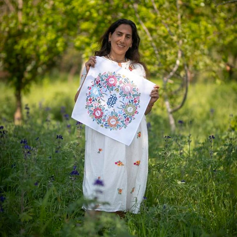 May include: A woman in a white dress holds a white fabric square with a colorful floral design and the word "Joy" in the center.