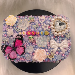 Puede incluir: Un objeto decorativo adornado con la palabra "LASHES" en cuentas de colores. El objeto está adornado con perlas, rosas, mariposas y una joya en forma de corazón, en tonos rosa, morado y blanco.