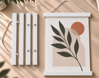 May include: A white wooden magnetic poster hanger frame holding a print featuring a dark green leafy branch, a light beige arch shape, and a solid terracotta circle. A second identical white wooden magnetic frame is shown disassembled nearby.