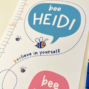 May include: A white height chart with a blue speech bubble that says "bee HEIDI" in white letters. The chart features a bee illustration and the phrase "bee-lieve in yourself". A measurement scale is visible on the left side, with markings for feet and centimeters.