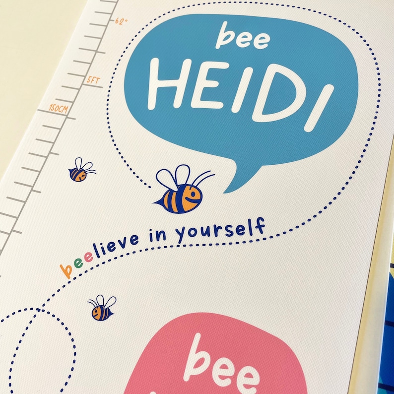 May include: A white height chart with a blue speech bubble that says "bee HEIDI" in white letters. The chart features a bee illustration and the phrase "bee-lieve in yourself". A measurement scale is visible on the left side, with markings for feet and centimeters.