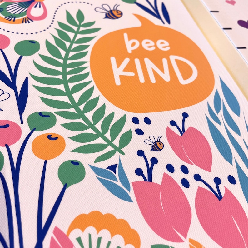 May include: A colorful floral print featuring the words "bee KIND" in an orange speech bubble. The design includes pink and blue flowers, green leaves, orange berries, and cartoon bees on a white background.