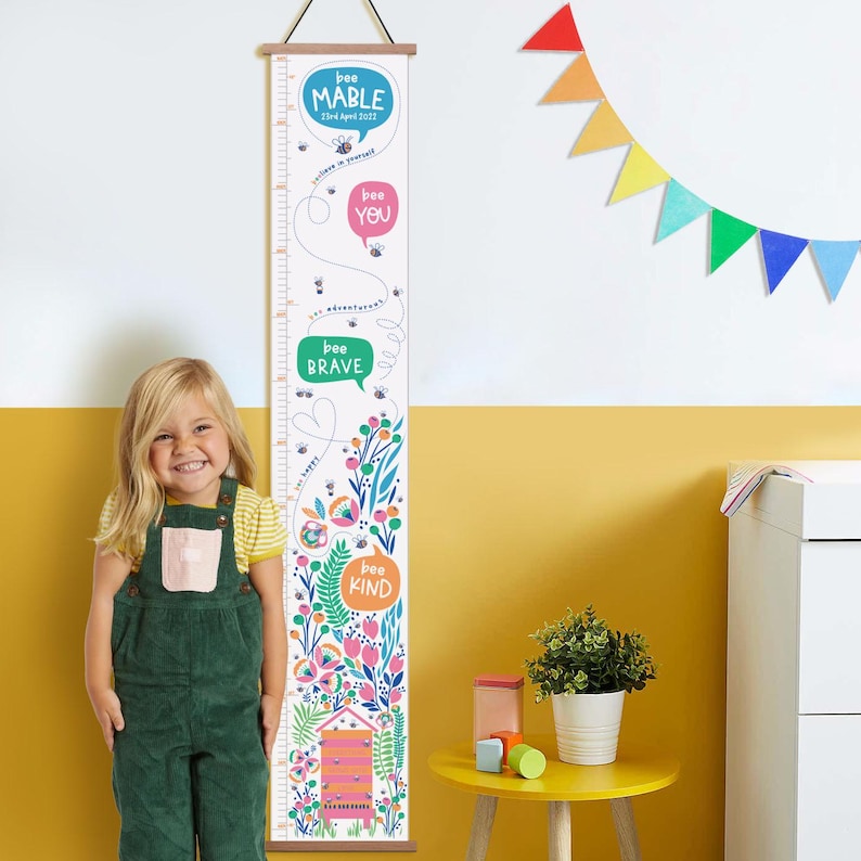 May include: A white height chart with a wooden top and bottom, featuring colorful floral and bee illustrations. The chart includes the phrases "bee mable", "bee you", "bee brave", and "bee kind", with a measurement scale in inches.