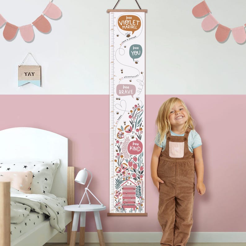 May include: A wooden height chart with a floral and bee design, featuring the phrases "bee Violet Maribel", "bee You", "bee Brave", and "bee Kind". The chart has a wooden top and bottom, and is designed to measure height in inches.