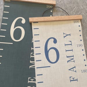 Two personalised canvas family growth charts with wooden frames. The chart on the left has a dark blue background with oak hangers. The family height chart on the right has a beige background with blue numbers and oak hangers. Both charts have a black string for hanging.