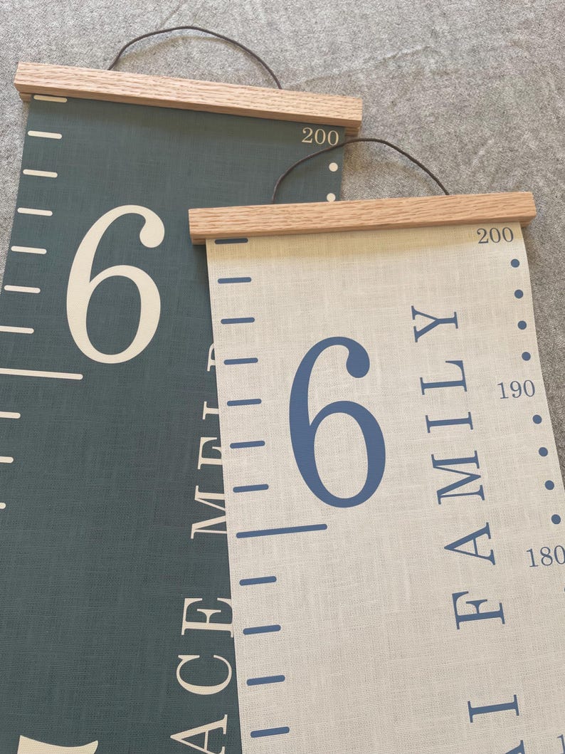 Two personalised canvas family growth charts with wooden frames. The chart on the left has a dark blue background with oak hangers. The family height chart on the right has a beige background with blue numbers and oak hangers. Both charts have a black string for hanging.