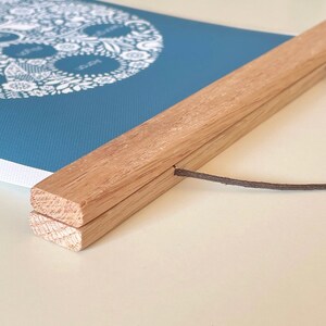 May include: A blue wall hanging featuring a white circular floral design with text, secured between two light-colored wooden slats. A thin brown cord is attached to the top wooden slat for hanging, providing a clean and simple display for the print.