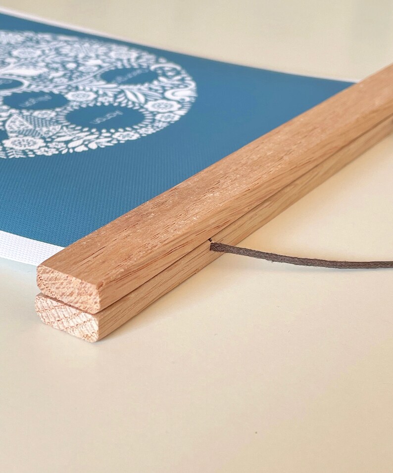 May include: A blue wall hanging featuring a white circular floral design with text, secured between two light-colored wooden slats. A thin brown cord is attached to the top wooden slat for hanging, providing a clean and simple display for the print.