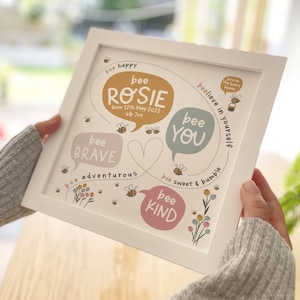 May include: White framed print with the name "Rosie" in a speech bubble, surrounded by bee illustrations and phrases like "bee brave" and "bee kind." The print includes the birth date of May 12th, 2022, and the weight of 6lb 7oz.