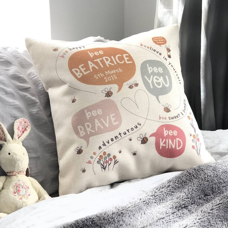 May include: A square, off-white decorative pillow with a bee-themed design. The pillow features speech bubbles with phrases like "bee brave" and "bee kind," along with the name "BEATRICE" and the date "5th March 2015".