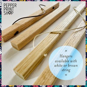 May include: Four light-colored wooden poster hanger bars with a visible grain. Two bars feature a dark brown leather-like cord, and two bars feature a light-colored string. Text reads: PEPPER PRINT SHOP and Hangers available with white or brown string.