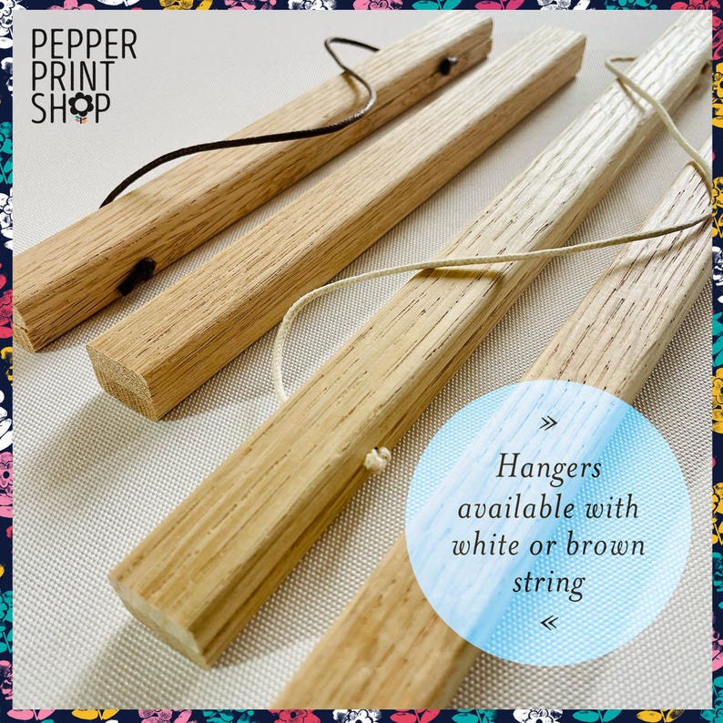 May include: Four light-colored wooden poster hanger bars with a visible grain. Two bars feature a dark brown leather-like cord, and two bars feature a light-colored string. Text reads: PEPPER PRINT SHOP and Hangers available with white or brown string.