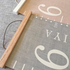Two personalised canvas family growth charts with wooden frames. The chart on the left has a light grey textured background with oak hangers. The family height chart on the right has a dusty pink background with white numbers and white hangers. Both charts have a string for hanging.