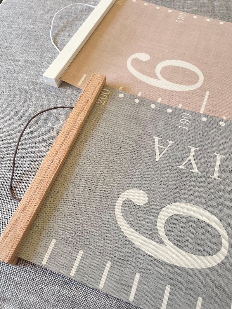 Two personalised canvas family growth charts with wooden frames. The chart on the left has a light grey textured background with oak hangers. The family height chart on the right has a dusty pink background with white numbers and white hangers. Both charts have a string for hanging.