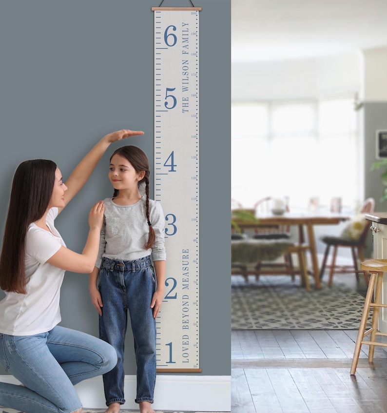 Pu&ograve; includere: Un grafico di crescita in tela bianca con testo blu che recita "Loved Beyond Measure" e "The Wilson Family". Il grafico &egrave; contrassegnato da numeri da 1 a 6. Un bambino &egrave; in piedi accanto al grafico, e un adulto lo aiuta a misurare la sua altezza.