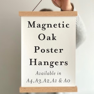 Pu&ograve; includere: Un supporto magnetico per poster in legno chiaro che sostiene un foglio rettangolare bianco con testo nero: Magnetic Oak Poster Hangers Available in A4, A3, A2, A1 & A0. Il supporto presenta un sottile cordino marrone sulla barra superiore.