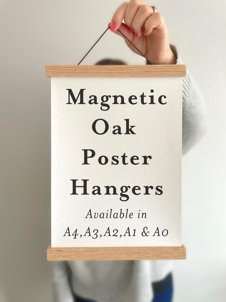 Pu&ograve; includere: Un supporto magnetico per poster in legno chiaro che sostiene un foglio rettangolare bianco con testo nero: Magnetic Oak Poster Hangers Available in A4, A3, A2, A1 & A0. Il supporto presenta un sottile cordino marrone sulla barra superiore.