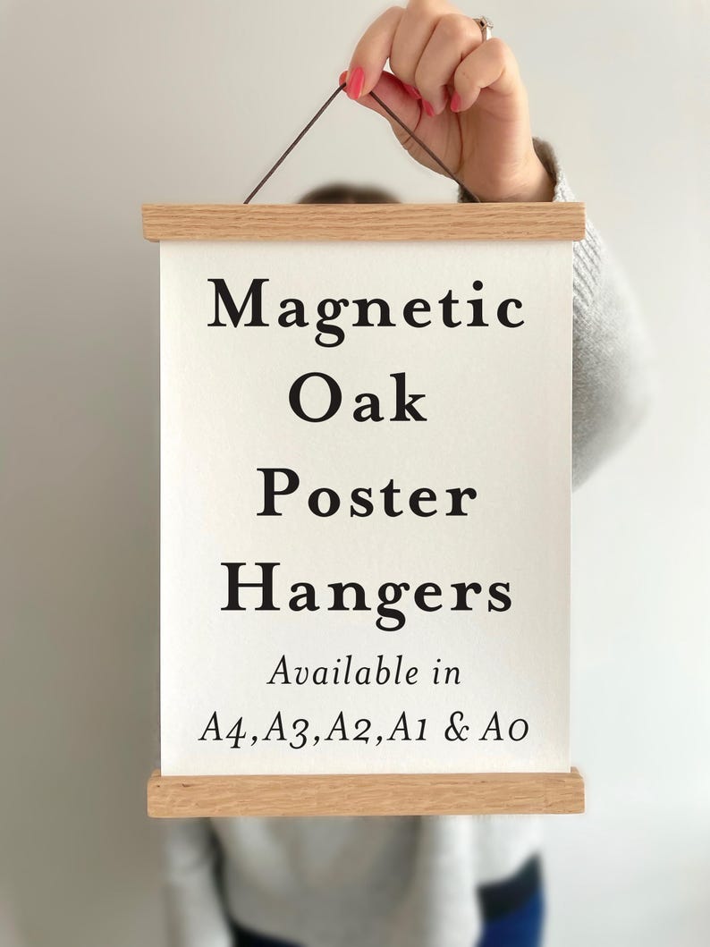 May include: A light-colored wooden magnetic poster hanger holding a white rectangular paper with black text that reads: Magnetic Oak Poster Hangers Available in A4, A3, A2, A1 & A0. The hanger features a thin dark cord for wall mounting.
