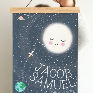 May include: A rectangular wall hanging featuring a dark blue space-themed print with a smiling moon, a rocket, a planet, and stars. The text reads JACOB SAMUEL and 18th April 2022. The print is secured between two light-colored wooden magnetic bars.