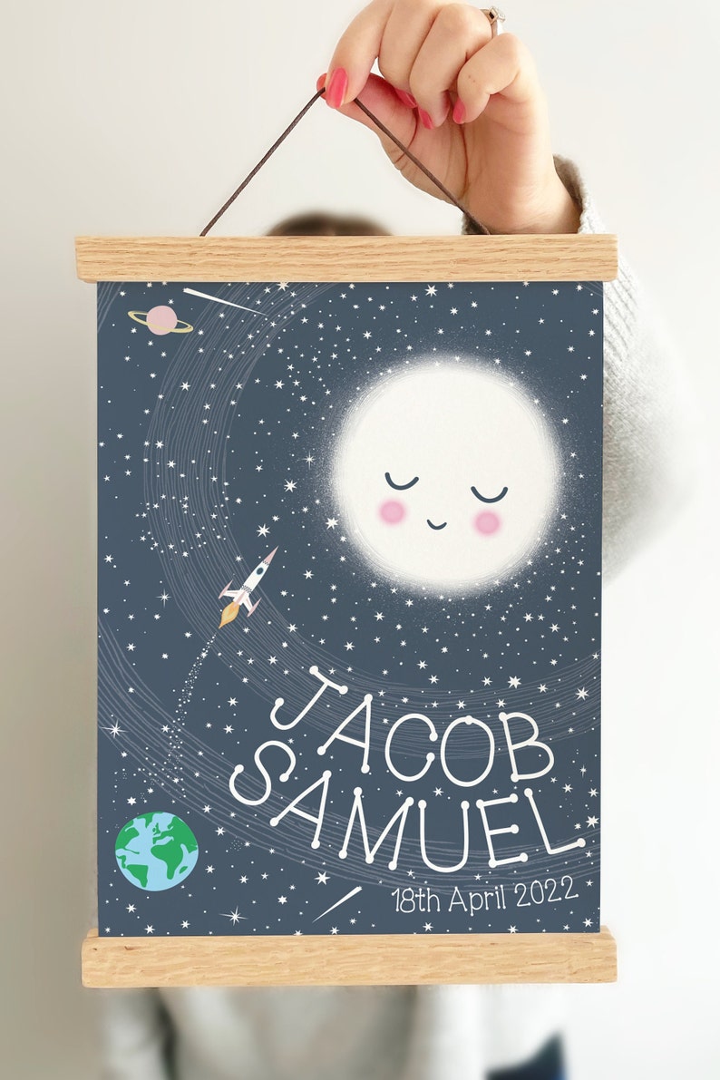 May include: A rectangular wall hanging featuring a dark blue space-themed print with a smiling moon, a rocket, a planet, and stars. The text reads JACOB SAMUEL and 18th April 2022. The print is secured between two light-colored wooden magnetic bars.