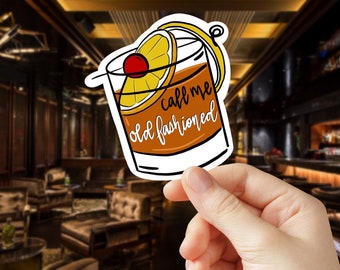 May include: A sticker featuring an Old Fashioned cocktail illustration. The drink is in a rocks glass with a cherry and orange slice garnish. Text on the glass reads "call me old fashioned" in a handwritten style. The sticker is held in front of a blurred bar background.