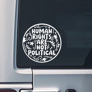 May include: White circular sticker with the text "HUMAN RIGHTS ARE NOT POLITICAL" in a decorative font. The text is surrounded by floral and leaf designs. The sticker is affixed to a vehicle window.