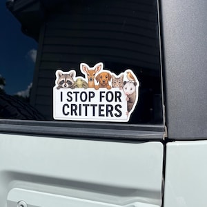 May include: A rectangular sticker with the text "I STOP FOR CRITTERS" and illustrations of a raccoon, turtle, deer, dog, cat, bird, and opossum. The sticker is affixed to a vehicle window.