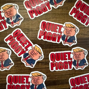 May include: A collection of stickers featuring a cartoon pig with blonde hair, wearing a suit and tie. The stickers have the text "QUIET, PIGGY" in red letters. The stickers are scattered on a wooden surface.