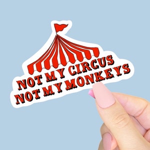 Puede incluir: Una pegatina blanca con un diseño de carpa de circo roja y blanca y el texto "NOT MY CIRCUS NOT MY MONKEYS" en letras rojas con contorno negro. La pegatina está sujeta por una mano con uñas rosas sobre un fondo azul claro.