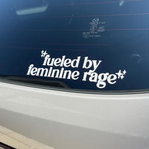 K&ouml;nnte beinhalten: Wei&szlig;er Vinyl-Aufkleber mit dem Text "#fueled by feminine rage" in fetter, serifenloser Schrift. Der Aufkleber ist auf der Heckscheibe eines Fahrzeugs angebracht, darunter ist die hellfarbene Karosserie zu sehen.