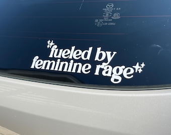 May include: White vinyl decal with the text "#fueled by feminine rage" in a bold, sans-serif font. The decal is affixed to the rear window of a vehicle, with a light-colored car body visible below.
