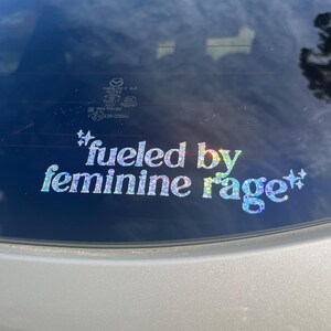 K&ouml;nnte beinhalten: Ein holografischer Aufkleber mit dem Text "fueled by feminine rage" in einer verspielten Schriftart. Der Aufkleber ist an der Heckscheibe eines silbernen Fahrzeugs angebracht und reflektiert den Himmel und die Umgebung.