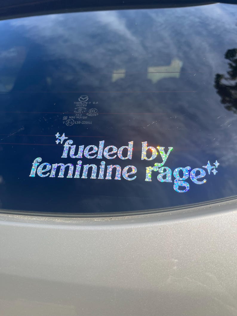 K&ouml;nnte beinhalten: Ein holografischer Aufkleber mit dem Text "fueled by feminine rage" in einer verspielten Schriftart. Der Aufkleber ist an der Heckscheibe eines silbernen Fahrzeugs angebracht und reflektiert den Himmel und die Umgebung.