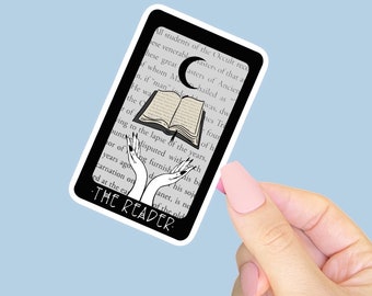 May include: A black and white sticker featuring a tarot card design. The card depicts an open book, a crescent moon, and stylized hands. The text "THE READER" is at the bottom. The sticker is held by a person with a light blue background.