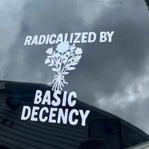May include: White sticker on a car window with the words "RADICALIZED BY" above a floral bouquet graphic and the words "BASIC DECENCY" below. The sticker is against a dark, cloudy background.