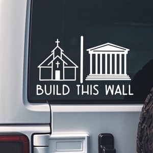 May include: White vinyl sticker on a silver car with a church and a building with columns. The text says "BUILD THIS WALL".