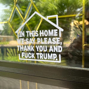May include: White vinyl decal in the shape of a house, affixed to a glass door. The text reads: "IN THIS HOME WE SAY PLEASE, THANK YOU AND FUCK TRUMP."