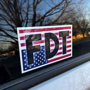 Puede incluir: Una pegatina con las letras "FDT" en negro sobre un fondo de bandera estadounidense roja, blanca y azul.