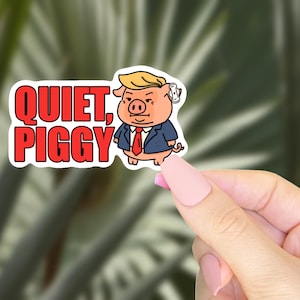 May include: A sticker featuring a cartoon pig wearing a suit and tie, with blonde hair and a small earpiece. The sticker has the words "QUIET, PIGGY" in red block letters. The sticker is held by a hand with pink nail polish.