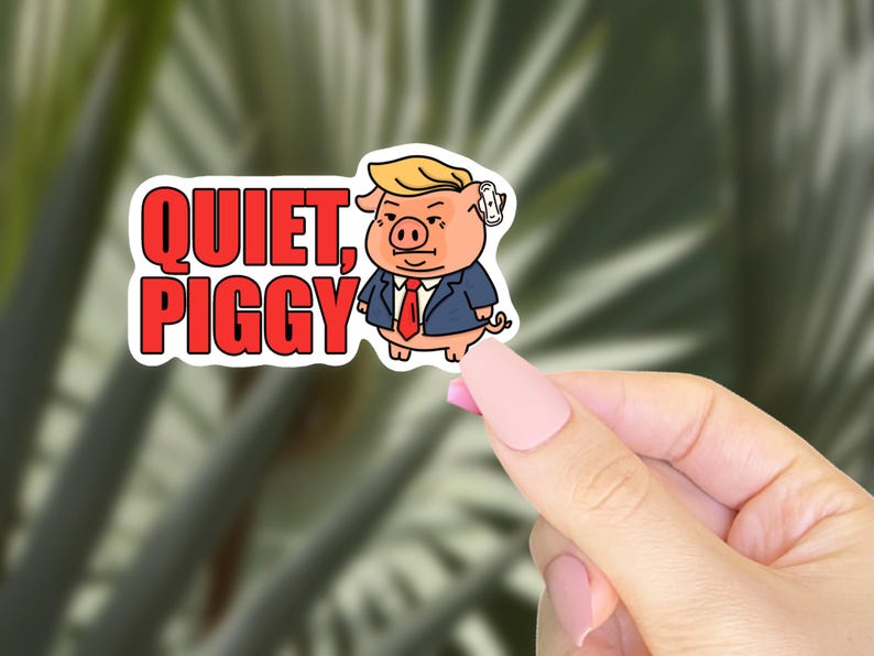 May include: A sticker featuring a cartoon pig wearing a suit and tie, with blonde hair and a small earpiece. The sticker has the words "QUIET, PIGGY" in red block letters. The sticker is held by a hand with pink nail polish.