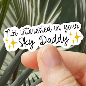 Puede incluir: Una pegatina blanca con el texto "Not interested in your Sky Daddy" en escritura negra. La pegatina tiene un borde negro y está decorada con dos estrellas amarillas. La pegatina está sujeta por los dedos de una persona.